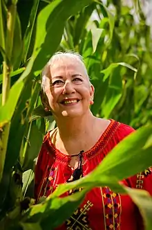 Portrait of a woman with short, white hair wearing a red, embroidered blouse tied near her sternum surrounded by tall green plants with broad leaves.