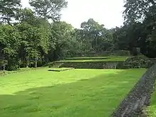 A flat grassy area with low stepped structures at the far side, bordered by trees