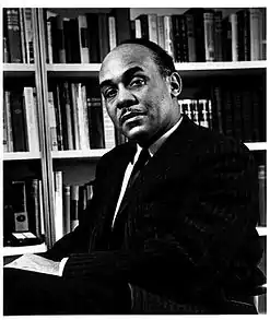 Ralph Ellison is pictured sitting in a chair before a bookcase. He is wearing a suit and has a mustache and receding hair-line.