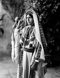 Image 7A young woman from Ramallah, c. 1898-1914.  Until the 1940s, women of Palestine wore elaborate handcrafted garments.  The creation and maintenance of these items played a significant role in their lives.  A knowledgeable observer could determine a woman's village of origin and social status from her clothing.  The circular band near this woman's forehead is a ring of coins made from a portion of her dowry money, and indicates that she is unmarried.'Ali Ra'if Efendi, (edited by Durova)