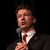 A man with brown, wavy hair wearing a suit and holding a microphone