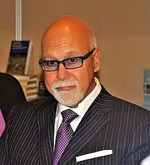 Photograph of René Angélil.  Depicting his head an upper torso, the man has a bald head, tanned skin, and grey goatee beard and moustache.  He is wearing spectacles, white shirt with purple necktie, and a pinstriped suit jacket.