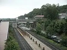 A low-level railway platform between two tracks. A parking lot is on the right. On the left, a road overpass rises to cross the tracks in the background.