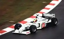 A figure, wearing a helmet of a dark blue design with red and yellow stripes, is driving a Formula One with an white colour scheme adorned with sponsorship. He is holding both hands on a steering wheel, turning right.