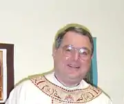 A bespectacled man wearing a white chasuble with beige and burgundy ornamentation around the neckline