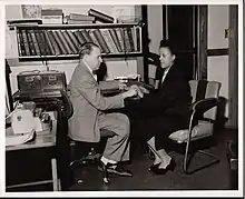 Black and white photo of two people sitting in an office. Kinney, a middle-aged white man dressed in a suit, has his hand in the palm of Lawhorn, a middle-aged Black woman wearing a dark-colored dress.