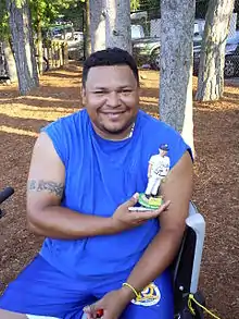 A man wearing a blue sleeveless T-shirt and blue shorts smiles while holding a bobblehead depicting himself in a white baseball uniform.