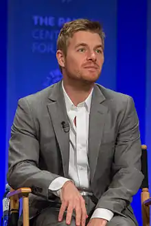 Man sitting in a chair with a blue wall behind him.