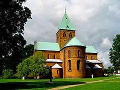 A cruciform, copper-roofed, brick church in Romanesque style with rounded window arches and domed apse. The large tower has pointed Gothic-styled windows.