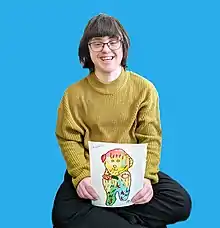 The image features a woman (Rita Winkler) sitting against a blue background, smiling broadly. She is wearing glasses with a rounded frame, a mustard yellow knit sweater, and black pants. The woman is holding a colorful painting of a whimsical character with a rounded head, decorated with patterns and stars. She has characteristics of Down syndrome such as almond-shaped eyes and a flat profile.