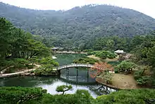 A pond with a bridge and islands in a park setting surrounded by wood and in front of a hill.