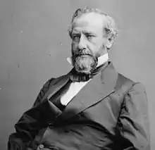 A man with receding, graying hair, a mustache, and a long beard wearing a dark jacket, vest, and tie and a white shirt