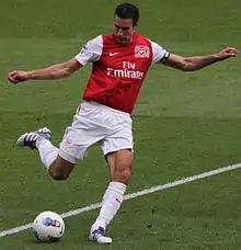 Robin van Persie, wearing a red and white football jersey and white shorts, prepares to kick a football with his right foot with both arms outstretched.