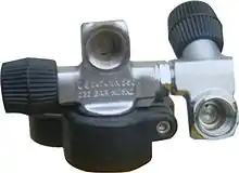  A valve body is shown in place on a cylinder neck, with a cylinder handle clamped below. The valve has a DIN connection socket in line with and perpendicular to the cylinder axis, and the orthogonal right hand operated main valve spindle has a plastic knob. Opposite this knob, and on the axis of this spindle, there is a socket into which a secondary valve body has been screwed, using left hand thread and a lock nut. This secondary valve also has a DIN connection socket, on a stub branch perpendicular to its inlet axis, orientated downwards and facing in the same direction as the main outlet, towards the viewer. The secondary valve knob is roughly opposite to the secondary outlet and the axis points upward and slightly backward. The main valve and spindle valve axes form the shape of a capital H.