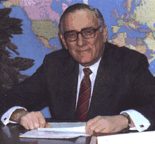 Middle-aged man with black and gray hair sitting behind a desk, wearing a navy blue suitcoat, white dress shirt, red necktie, and black-rimmed eyeglasses