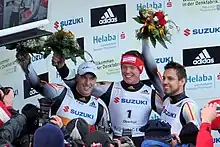 Three smiling men stand side-by-side in front of a crowd of photographers. Each holds up a flower bouquet with their right arm, and wears the same tight black-and-white jumpsuit with some yellow and red stripes. The man on the left wears a white cap, while the one in the middle wears a red headband.