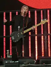 Roger Waters, dressed in black, playing a bass guitar and speaking into a microphone. Behind him are several red vertical video panels.