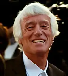 An older man with medium-length white hair and tanned skin smiles at the camera.