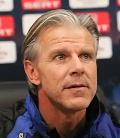 A middle-aged man of Nordic appearance photographed at a sporting press conference. His greying blond hair is brushed back, stubble is clearly visible on his face, and his dark blue eyes look up and to the viewer's right.