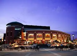 On the left is a two-storey red brick rotunda with a full-height five-pane wide dark window showing parts of the interior atrium, and at which base is the main entrance to the theatre. To the right is a red brick wall arcing away, with nine identical and equally spaced four-pane wide windows stretching from the ground to nearly the roof. It is fronted for its entire length by a stairway with about seven steps, leading to a piazza in the foreground.