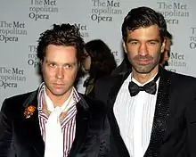 Two men wearing suits; the one on the left is wearing a red-and-white-striped dress shirt along with a floral brooch and white scarf, while the one on the right is wearing a black bow-tie. The text "The Metropolitan Opera" is repeated in the background.