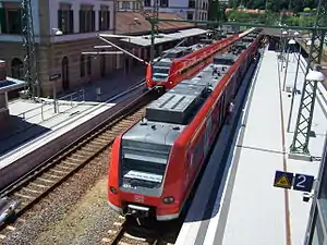 Pair of red electrical-multiple-unit passenger trains running from overhead-catenary power called at a railway station with at least four tracks and three platforms, two of which are on an island and one of which is to the side directly connected to the main station building, with all platforms having sheltered and unsheltered sections