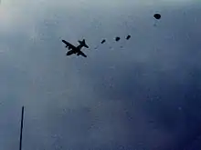 Four small parachutes can be seen coming out the back of a distant, silhouetted plane in flight. It is daytime, however the sky is dark and cloudy.