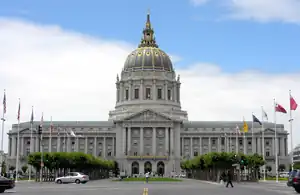 A large three story building with a wing on either side of a tower. The building is white, with a blue and gold dome on top of the tower. In front of the building is a plaza lined with flagpoles and trees.