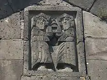 Two bearded figures in stone relief, holding an object that looks like an open door with a bell on top.