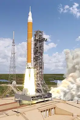  A large orange and white rocket launching off pad 39B at Kennedy Space Center beside a tall steel support tower