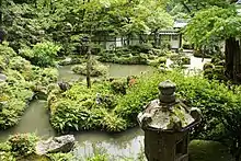 A color photo of a Japanese landscaped garden with mounds of greenery, trees, rocks, a pond and, in the foreground, a stone lantern (dominant color: green)