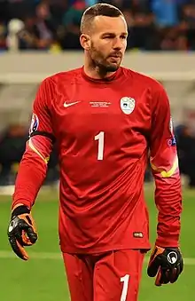 A dark-haired white man, wearing an all red football kit and black goalkeeping gloves.