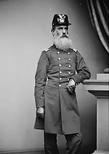 An 1862ish black and white photograph of a white male army cavalry officer with a long full white beard age 60 in full military uniform wearing an older style dragoon headwear.