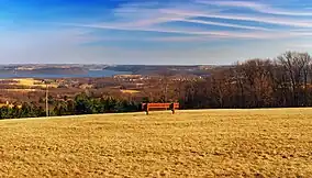 A red bench on a large expanse of brown cut grass, overlooking rolling forested hills and a lake