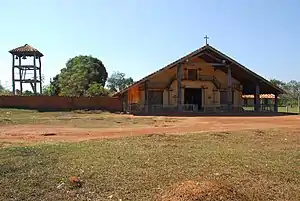 A wooden bell-tower and a church from in frontal view beyond a grassy area. A cross is positioned at the top of the roof.