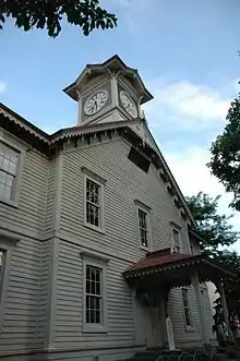 Photograph of a white wooden building against a blue sky.  The building is of Western architectural design built with subtle Japanese details.  A clock tower rising from the front of the building is the dominant feature.