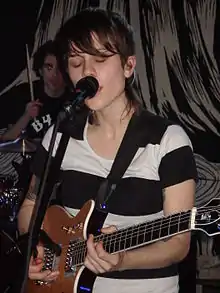 Sara Quin performing with a guitar and singing into a microphone with a drummer in the background