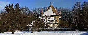 A snow covered field and dark trees.  In the center the square castle building with three visible corner towers all of large rectangular brown stone.  The roof is sharply peaked, of red-brown tile.  Outbuildings partially block the view of the castle and are white with red roofs.