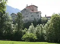 A light grey, compact, square castle with a low square tower in the center, surrounded by green trees and a hazy sky.