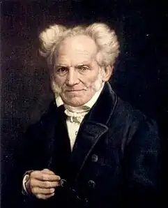 A portrait of a man wearing a black coat, looking straight ahead, unsmiling, holding his right hand in front of himself. He is bald, and the white hair on the sides of his head are pointing outward.