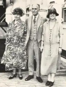  Three white people stand on a ship's deck for a photo: a older woman in a floral dress and dark hat, an older balding man in a suit, and a young woman in a cloche hat and dress with dark buttons down the front