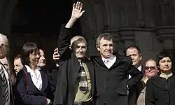 Two hugging men stand in a small crowd facing slightly to the right. The man on the left has his right hand raised to the crowd behind the camera, and wears glasses, a black suit and olive green shirt. The man standing next to him has a black suit, white shirt and pink tie. He has his right arm on the left man's right shoulder and his left arm on his own hip.