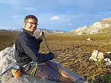 Image of Sean Dougherty in climbing gear. He is sitting on a rock on the left side of the image, smiling to the camera, while wearing technical climbing gear and handling a climbing rope.