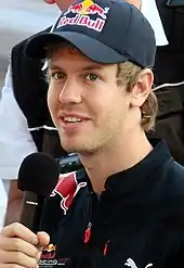 Sebastian Vettel wearing a sponsored baseball cap and holding a black microphone in his right hand