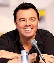 A man with black short hair and a black shirt, with tan skin, laughs into a microphone while leaning forward.