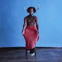 A photo of the artist sat on a stool in the middle of a room with blue walls and a dingy concrete floor.