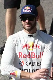 A man sporting a beard in his late twenties is wearing sunglasses and a dark blue baseball cap with a sponsor logo. His right hand is on his left arm.