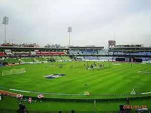 Front view of the entrance and a stand of a cricket stadium. Some people can be seen in the ground.