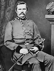 Black-and-white photo of a mustachioed military officer sitting with a saber across his lap