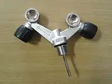  a parallel thread cylinder valve is shown, with a chromed dip tube, and two DIN valved connection sockets. The sockets are offset from the angled arms of the valve body, which branch at roughly 45 degrees from the neck thread axis in the same plane. The connection socket axes are orthogonal to the plane of the arms, and the valve spindles are orthogonal to their outlets and the axis of each arm, with the knobs to the outsides.